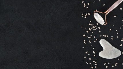 Home spa salon with gouache scraper and facial massage roller. Trending skin care. Lifting for healthy and youthful skin. Organic cosmetics on dark background with pink sea salt. Copy space. Top view.