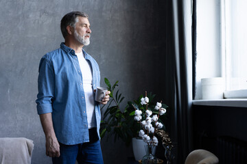 Handsome senior man is holding a cup, looking away and smiling while standing in apartment.