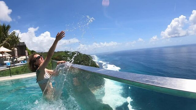 Caucasian woman splashes water in the infinity swimming pool overlooking an ocean.Camera moves along with the splash 