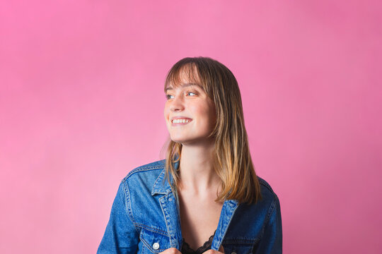 Studio portrait of a woman, she is listening to music and is smiling, happy and full of emotions. Standing in the center of the image, in front of a pink background.