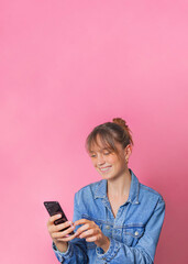 Studio portrait of a woman holding a phone, she is texting and she is happy. She is smiling and full of emotions. She is standing in the center of the image, in front of a pink background.