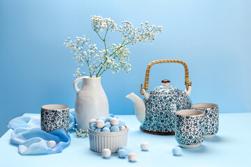 Teapot in a blue flower, cups, white vase with flowers and round caramel candies on a blue background. Blue still life with utensils. High quality photo - Powered by Adobe
