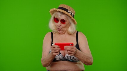 Senior woman tourist in swimsuit bra playing video game on mobile cell phone, using mobile app drive simulator, spending leisure time on internet entertainment on chroma key background. Exited granny