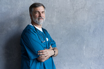 Portrait of professional mature surgeon looking at camera while standing against the grey background.