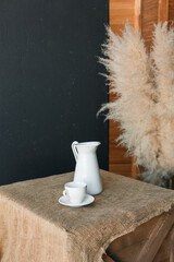 White decanter, white cup on white saucer, stand on burlap bedding on blue textured background and dry reed branches