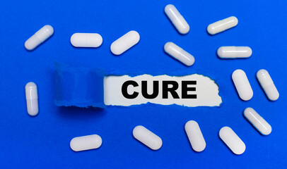 White pills lie on a beautiful blue background. In the center is white paper with the inscription CURE. Medical concept. View from above.