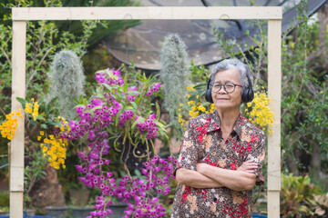 Cheerful senior woman with short gray hair, wearing wireless headphones, smiling, arms crossed and looking at the camera while standing in a garden. Concept of aged people and relaxation