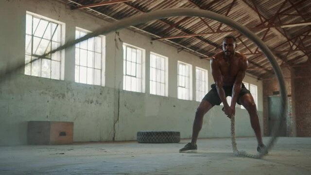 Athletic young man with battle rope doing exercise in training gym. Shirtless man working out with battle ropes inside abandoned warehouse.
