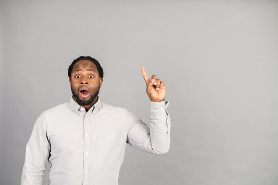 Excited African-American guy came up with a new solution, a biracial surprised man wearing smart casual shirt points finger up, empty copy space for advertising text, isolated on grey