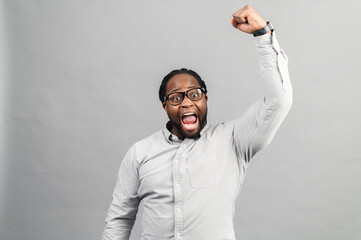 Excited African-American guy wearing smart casual shirt celebrating victory, raising clenched fist up, screams yes happily, black guy yelling ecstatic, triumph concept, studio shot isolated on grey