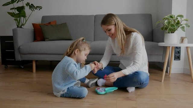 Mother and little daughter paint their nails with toy nail polish. Kid playing with mom at home. Concept of good parenting and happy childhood, family leisure