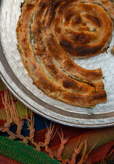 homemade Bosnian pie burek with meat in a traditionally made vessel and old homemade Persian style rug and wooden plate