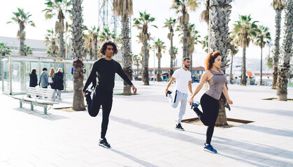 Obraz premium Attractive female fitness instructor in electronic headphones showing exercising method to ethletic clients enjoying morning workout, group of diverse people collaborating together on stretching