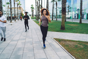 Cheerful curly woman in active wear enjoying running on cardio training in city slimming and losing weight, happy beautiful female jogger in trendy sportswear satisfied with body physical strength