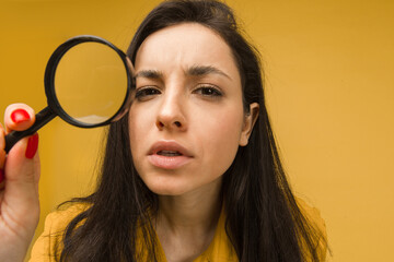 Photo of cute female holds magnifying glass and looking for something. Wears yellow shirt, isolated yellow color background