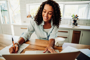 Mixed race female working from home writing in note pad studying for tests typing on laptop