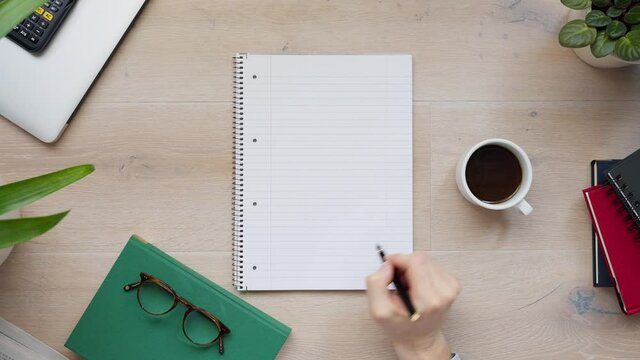 Top view shot of a blank page of notebook with a person pretending to write with his black fountain pen. Calculator, glasses and books on light wooden table. 4k Close-up daylight