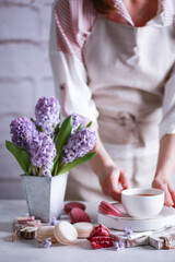 Spring atmosphere. Flowers in a vase, macaroni, chocolate and a cup of tea on wooden white boards on a light table with hands. Lilac hyacinths. Background image, copy space, vertical, rustic