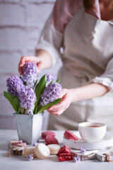 Spring atmosphere. Flowers in a vase, macaroni, chocolate and a cup of tea on wooden white boards on a light table with hands. Lilac hyacinths. Background image, copy space, vertical, rustic