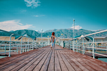 Red-haired girl runs along the pier. Woman in flowing long dress. Marine wooden pier with umbrellas. Pretty woman is enjoying vacation. Mountains in the background. Rear view. Hot summer day.