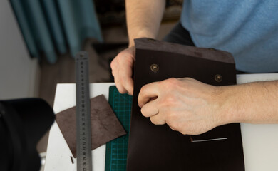 Mens hand working on the leather wallet in his workshop. Working process with a brown natural leather. Craftsman holding a crafting tools.