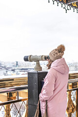 A young girl looks through coin-operated binoculars on the observation deck overlooking the city from a height at sunset. Winter, snowfall