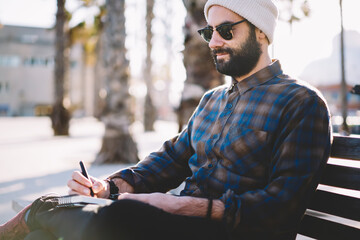 Middle Eastern male author in stylish sunglasses and hat writing text ideas in education notepad, creative hipster guy making informative notes while planning touristic sightseeing rest on bench