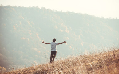 A man stands against the backdrop of a mountain landscape with his hands raised high. Autumn, sun, dawn.