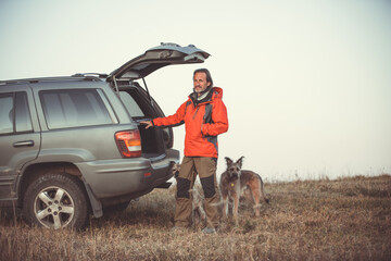 An adult man stands near a car with two dogs against the background of the sky. Traveling with pets.