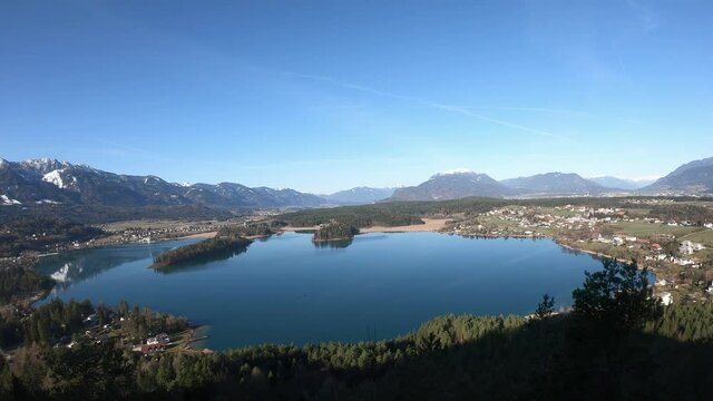 A panoramic view from a small mountain on Faak lake in Austrian Alps. The picturesque lake is surrounded by high mountains. The mountains are reflecting in calm surface of the lake. Lots of greenery
