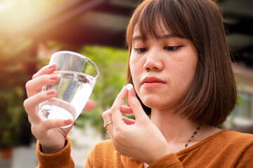The woman who is sick with fever is sick, holding a glass of water in the right hand. Left hand holding a pill with a blurred background.