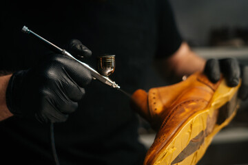 Close-up hands of unrecognizable shoemaker wearing black gloves spraying paint of light brown leather shoes. Concept of cobbler artisan repairing and restoration work in shoe repair shop.