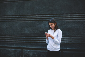 Cheerful young woman messaging via cellphone while standing near dark wall