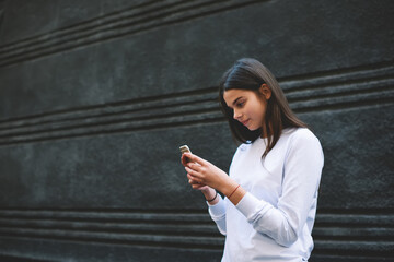 Focused millennial lady browsing smartphone while standing near concrete wall