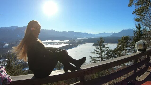 A woman in black sitting on a wooden rail and flipping her long hair with the panoramic view on Faak lake in Austrian Alps. The picturesque lake is surrounded by high mountains. Sun is shining bright.