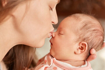 Mom with newborn baby posing indoor, mother kissing her sleepy infant, female closed eyes and pout lips, enjoying spending time with her child, motherhood.