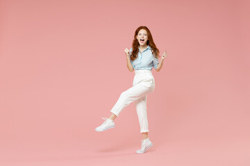 Fototapeta na wymiar Full length body fun young student lucky overjoyed excited happy redhead woman 20s in blue shirt pants walk do winner gesture clench fist celebrate isolated on pastel pink background studio portrait