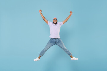 Fototapeta na wymiar Full length young overjoyed excited happy cheerful unshaven black african man in violet t-shirt hat glasses jump high with outstretched legs hands isolated on pastel blue background studio portrait.