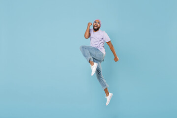Fototapeta na wymiar Full length young overjoyed unshaven black african man in violet t-shirt hat glasses do winner gesture jump high clench fist celebrate scream yes isolated on pastel blue background studio portrait