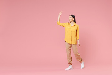Full length of young smiling happy brunette positive attractive latin woman 20s in yellow casual shirt walk going waving hand greeting someone isolated on pastel pink color background studio portrait.