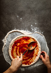 Making a pizza spreding tomato sauce on the dough on the grey concrete background - obrazy, fototapety, plakaty