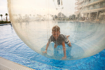Little boy have fun inside big plastic balloon on the water of swimming pool on the summer resort. Kid inside big inflatable transparent ball running and having fun.