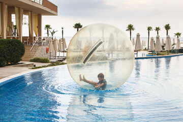Little boy have fun inside big plastic balloon on the water of swimming pool on the summer resort. Kid inside big inflatable transparent ball running and having fun. - obrazy, fototapety, plakaty