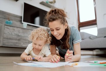 Smiling mother with her preschool-aged daughter spending their free time drawing together while lying on the wooden floor in the living room at home. Mom and daughter drawing with markers.