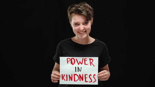 Positive laughing woman posing with Power in kindness poster at black background. Happy Caucasian female activist with toothy smile looking at camera smiling. Happiness and lifestyle.