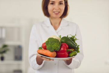 Close up of a plate of fresh vegetables that a young female nutritionist is holding in front of her. Concept of a healthy lifestyle, body care and weight loss. Selective focus. Blurred background.