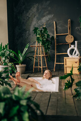 Vertical image of beautiful blonde young woman taking bath with bubbles in bathroom interior with plenty green plans