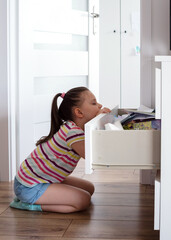 A little girl is looking for something in the chest of drawers among papers and drawings. A preschool girl tidies up the dresser in her room.