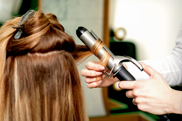 Fototapeta premium Back view of female hairdresser's hands curling women's hair with curling iron in a hair salon