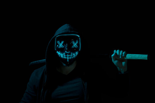 Man armed holding a katana and wearing a scary lighting neon glow mask and a hoodie on black background.